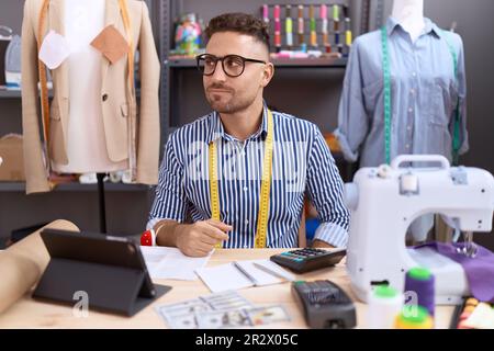 Homme hispanique avec couturier de la barbe designer travaillant à l'atelier sourire à la recherche du côté et regarder loin de penser. Banque D'Images