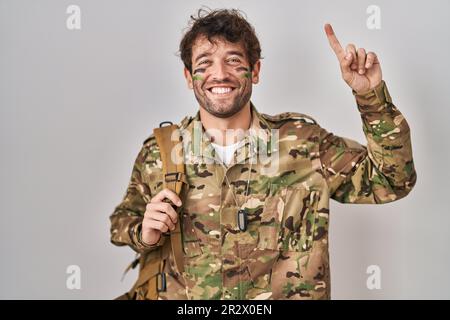 Un jeune homme hispanique portant un uniforme de l'armée de camouflage souriant surpris et surpris et pointant vers le haut avec les doigts et les bras levés. Banque D'Images