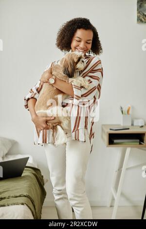 Portrait vertical de la jeune femme noire tenant le petit chien à la maison et souriant heureux Banque D'Images