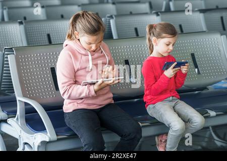 Enfants, enfants, filles attendant un vol en avion dans le hall des départs. Assis sur des chaises, jouer à des jeux d'ordinateur dans des gadgets, téléphone mobile, tablette. Discussion Banque D'Images