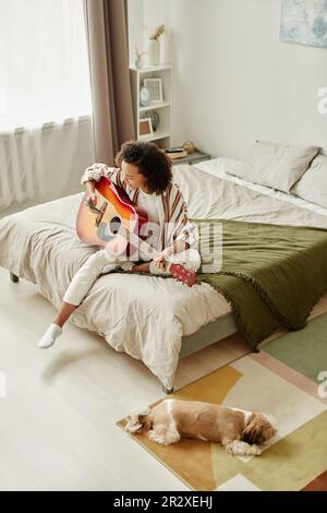 Portrait grand angle aéré d'une jeune femme jouant de la guitare acoustique sur le lit tout en se relaxant dans la maison confortable avec un chien d'animal de compagnie Banque D'Images