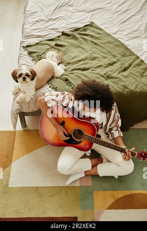 Vue de dessus de la jeune fille noire jouant de la guitare acoustique et se détendre à la maison avec un petit chien d'animal de compagnie Banque D'Images