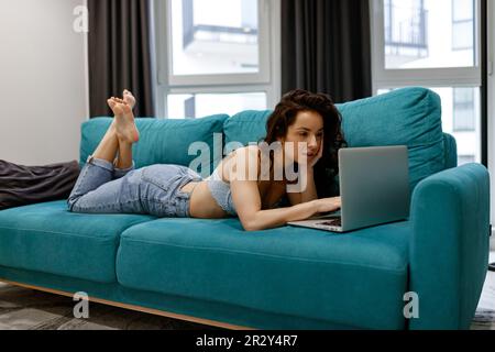 Belle jeune femme concentrée à cheveux foncés lisant des nouvelles sur l'écran d'ordinateur portable tout en se reposant sur le canapé à la maison. Concept de travail et de loisirs Banque D'Images
