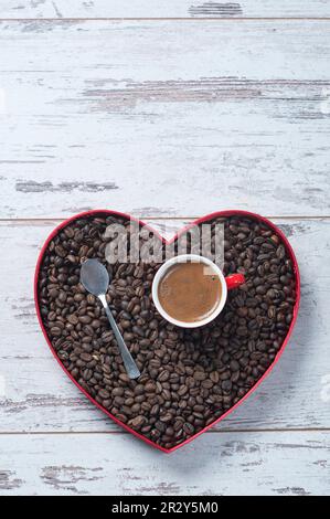 Vue de dessus d'une tasse d'espresso dans une tasse rouge sur des grains de café fraîchement torréfiés dans un plateau en forme de coeur sur une planche en bois blanc texturé Banque D'Images