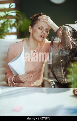 Un lieu de travail durable. femme moderne d'âge moyen employée dans un bureau moderne vert avec des documents et un ventilateur électrique souffrant de la chaleur estivale. Banque D'Images