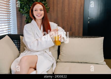 Portrait d'une jeune femme gaie portant un peignoir blanc en prenant un cocktail assis sur un canapé près de la fenêtre au spa. Charmante femme rouge Banque D'Images