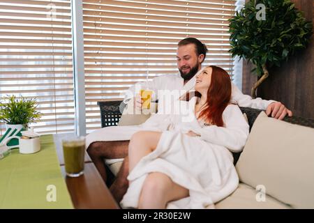 Portrait de jeune couple gai amoureux portant des peignoirs blancs, buvant des cocktails de fruits frais, parlant, se détendant assis confortablement Banque D'Images