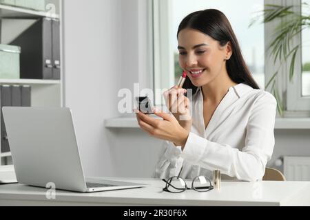 Jeune femme utilisant un miroir de poche cosmétique tout en appliquant le maquillage dans le bureau Banque D'Images