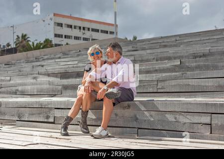 Un heureux couple d'âge moyen sourit en s'asseyant dans un escalier. Il est grizzli, avec une barbe blanche et portant des lunettes de soleil. Elle sourit et regarde le Banque D'Images