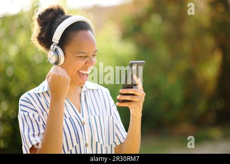 Femme noire excitée portant un casque célébrant l'écoute de musique avec le téléphone dans un parc Banque D'Images