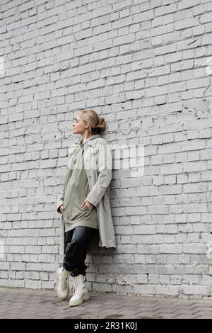 jeune femme à maquillage, cheveux blonds, langes, dans une tenue élégante avec long sweat à capuche, manteau, pantalon en cuir noir et bottes beiges debout avec les yeux fermés n Banque D'Images