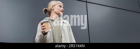 jeune femme tendance aux cheveux blonds avec des lanières debout dans un manteau et un sweat à capuche tout en tenant une tasse en papier avec un café à emporter près du bâtiment gris moderne Banque D'Images