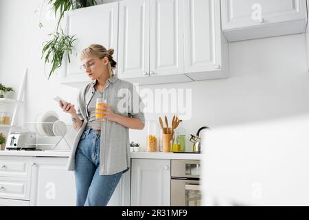 femme tatouée avec des langes et des lunettes tenant un verre de jus d'orange et utilisant un smartphone tout en se tenant près de plats propres et de plantes vertes dans le blurr Banque D'Images