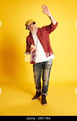 Portrait complet d'un homme à barbe mûr dans des vêtements élégants, une chemise à carreaux et un chapeau, posé avec un café sur fond jaune de studio Banque D'Images