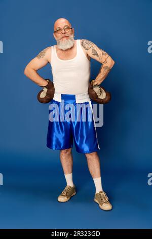 Portrait d'homme à tête blanche, barbu, mature en vêtements de sport et lunettes, avec corps tatoué en gants de boxe vintage sur fond bleu studio Banque D'Images