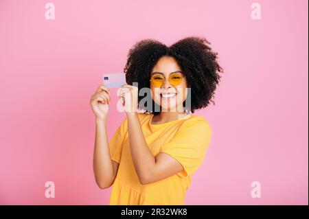 Photo d'une charmante femme brésilienne ou hispanique aux cheveux maureux avec des lunettes jaunes, vêtue de robe jaune, tenant les mains carte bancaire, debout sur un fond rose isolé, regarde l'appareil photo, sourit Banque D'Images