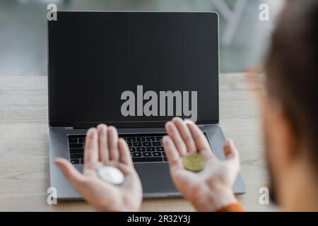 vue rognée d'un homme d'affaires flou tenant des bitcoins sur les paumes ouvertes tout en étant assis près d'un ordinateur portable avec écran blanc sur le bureau moderne, blu Banque D'Images