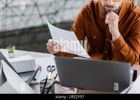 vue rognée d'un homme d'affaires attentif et occupé tenant un document près d'un ordinateur portable, de dossiers, de ciseaux, d'un marqueur et d'un porte-stylo avec des stylos pendant le travail Banque D'Images