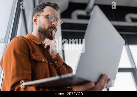 vue en contre-plongée d'un homme d'affaires à barbe sérieux avec lunettes et chemise tendance, regardant un ordinateur portable et pensant tout en étant debout au bureau sur une perte floue Banque D'Images