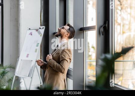 vue latérale d'un homme d'affaires plein de joie dans un blazer beige élégant et des lunettes penchées sur les fenêtres et riant en se tenant près du tableau de conférence avec n collant Banque D'Images