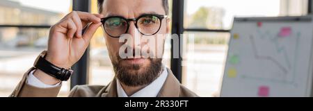 portrait d'un directeur d'entreprise barbu confiant et réussi en tenue officielle et montre-bracelet regardant l'appareil photo et ajuster les lunettes tendance en offi Banque D'Images