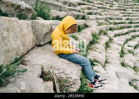 Enfant d'école jouant avec son téléphone portable smartphone dans un ancien amphithéâtre en pierre. Garçon à capuchon jaune en envoyant un SMS au-dessus de son gadget de téléphone pendant Banque D'Images