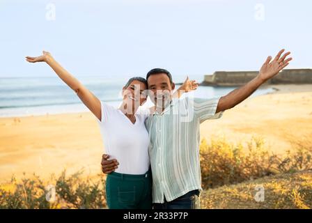 Joyeux couple d'âge mûr levant les mains posant près d'Ocean Beach en plein air Banque D'Images