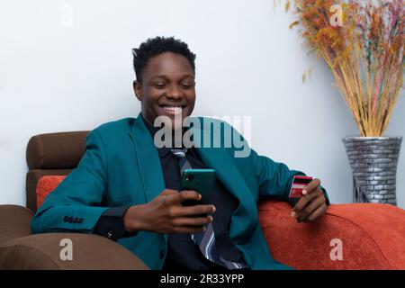 beau jeune homme noir portant un costume à l'aide de son téléphone et de sa carte de crédit, assis sur une chaise Banque D'Images