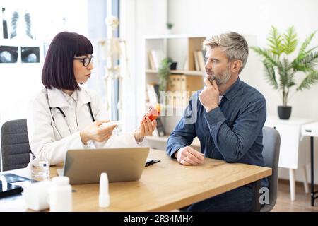 Femme médecin fournissant un examen expert et donnant des médicaments aux patients d'âge moyen dans le bureau de l'hôpital. Homme concentré écoutant attentivement les recommandations concernant les antécédents médicaux conservés sur ordinateur portable. Banque D'Images