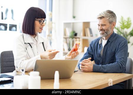 Femme médecin fournissant un examen expert et donnant des médicaments aux patients d'âge moyen dans le bureau de l'hôpital. Homme concentré écoutant attentivement les recommandations concernant les antécédents médicaux conservés sur ordinateur portable. Banque D'Images