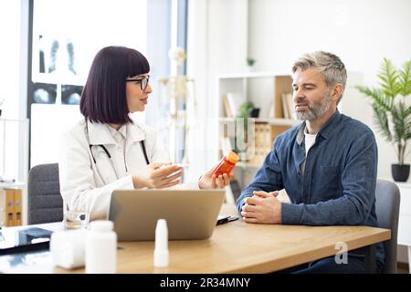 Femme médecin fournissant un examen expert et donnant des médicaments aux patients d'âge moyen dans le bureau de l'hôpital. Homme concentré écoutant attentivement les recommandations concernant les antécédents médicaux conservés sur ordinateur portable. Banque D'Images