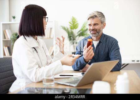 Vêtu d'un uniforme blanc, une femme thérapeute est équipée d'un ordinateur portable sur la table et consulte un patient mature lors d'une réunion de contrôle au bureau de l'hôpital. Homme souriant gardant une bouteille de pilules et à l'écoute des conseils du médecin. Banque D'Images