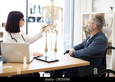 Jolie femme sous le manteau du médecin pointant vers le squelette humain tandis que le mâle mûr est assis en fauteuil roulant au bureau dans la salle de consultation. Illustrateur d'intention Banque D'Images