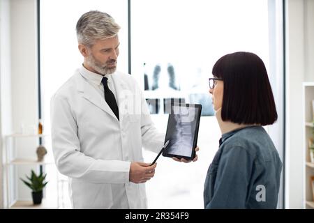 Beau gris-cheveux mâle en manteau blanc pointant vers le comprimé tandis que jolie femme dans le col médical restant dans la salle de conseil. Expert en soins de santé analysant des radiographies des vertèbres cervicales à l'intérieur. Banque D'Images