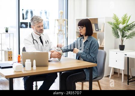 Homme caucasien mature en blouse de laboratoire mettant un appareil électronique sur le poignet de la femme pendant le contrôle dans la salle de conseil. Généraliste appliquant un oxymètre de pouls à une patiente en clinique médicale. Banque D'Images