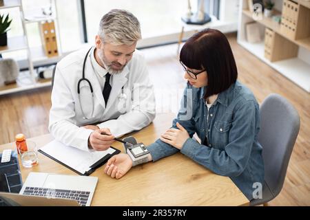 Vue de dessus de l'adulte barbu avec stéthoscope et brunette en denim tenue surveillant le tonomètre électronique sur le poignet. Médecin gai analysant les résultats positifs suivis par appareil au bureau du médecin. Banque D'Images