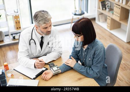 Vue de dessus de l'adulte barbu avec stéthoscope et brunette en denim tenue surveillant le tonomètre électronique sur le poignet. Médecin gai analysant les résultats positifs suivis par appareil au bureau du médecin. Banque D'Images