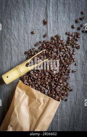 Les grains de café se répandre à partir d'un sac de papier avec un bambou scoop sur un tissu en lin gris Banque D'Images