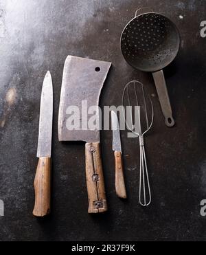 Vieux ustensiles de cuisine sur un fond sombre : cale de viande, cuillère en fil métallique, couteaux et passoire Banque D'Images