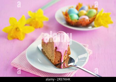 Un morceau de gugelhupf avec un glaçage de sucre rose, un nid de caramel avec des oeufs de sucre colorés et des jonquilles en arrière-plan Banque D'Images