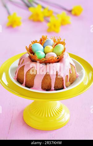 Un gugelhupf avec un glaçage au sucre rose et un nid au caramel avec des œufs de sucre colorés sur un stand, avec des jonquilles en arrière-plan Banque D'Images