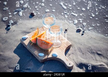 Deux verres de vin blanc et des tranches de melon de cantaloup sur une plage de sable Banque D'Images