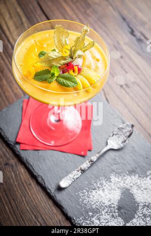 Sorbet mangue décoré de zeste d'agrumes, de menthe, de groseille dans un verre avec une serviette rouge et une cuillère sur un panneau d'ardoise et un fond en bois Banque D'Images