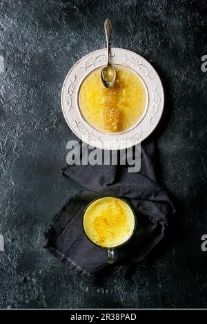 Nid d'abeille en miel liquide sur plaque vintage blanche et tasse de latte curcuma sur toile sur fond de texture noire Banque D'Images