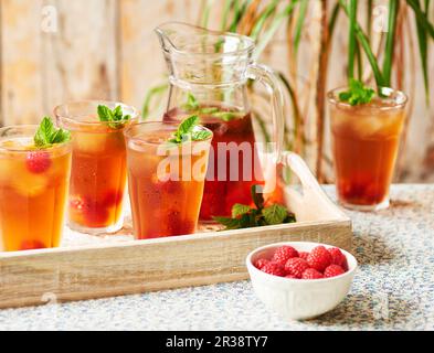 Une carafe et des verres de thé glacé Rooibos servis avec des framboises et de la menthe sur un plateau en bois posé sur une nappe florale Banque D'Images