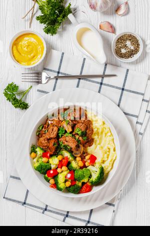 bœuf haché, ragoût de champignons servi avec une purée de pommes de terre et de pois chiches, tomates, brocolis, salade de fleurs dans un bol blanc sur une table en bois blanc avec de la musta Banque D'Images