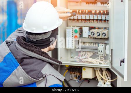 Un électricien professionnel connecte les fils dans la salle des panneaux électriques à l'extérieur. Le travailleur en casque de sécurité et en vêtements d'hiver vérifie l'équipement à l'aide d'un tournevis. Vue arrière. Banque D'Images