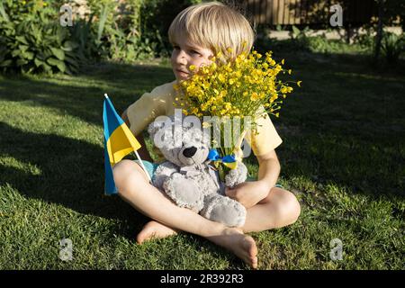 Malheureux garçon de sept ans assis sur l'herbe avec l'ours en peluche, drapeau ukrainien, bouquet de fleurs jaunes. Enfants contre la guerre.concept de paix pour l'Ukraine Banque D'Images