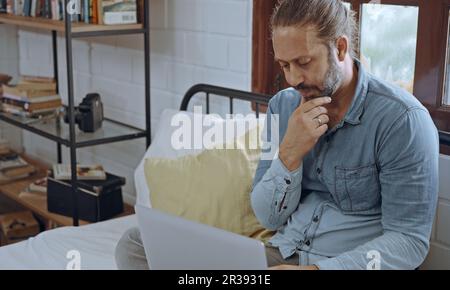 Un jeune homme inquiet à l'aide d'un ordinateur portable assis sur le lit dans la chambre. Banque D'Images
