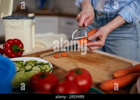 Femme préparant une salade végétarienne à la maison. Une femelle épluche une carotte avec un outil de pileur et recycle les restes dans un bac à compost. Durable Banque D'Images